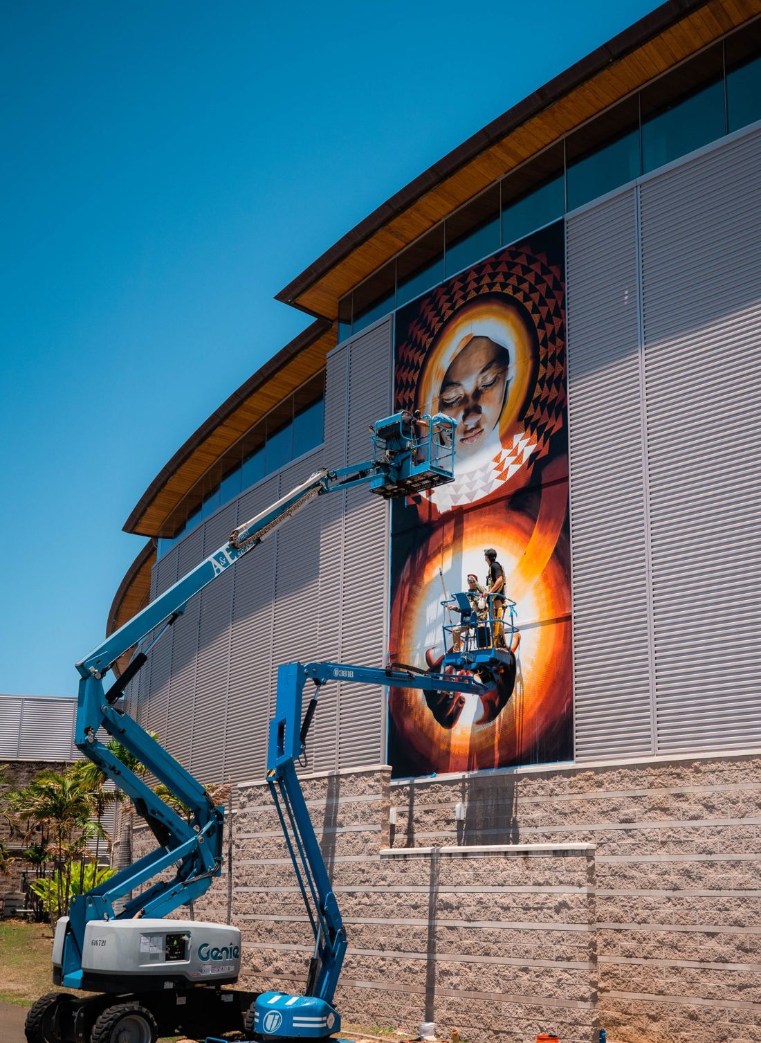 An artist on a blue boom lift painting a tall mural on the side of a museum building.
