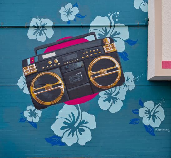 Mural on a blue brick wall of a retro boombox over a pink circle, surrounded by light blue hibiscus flowers.