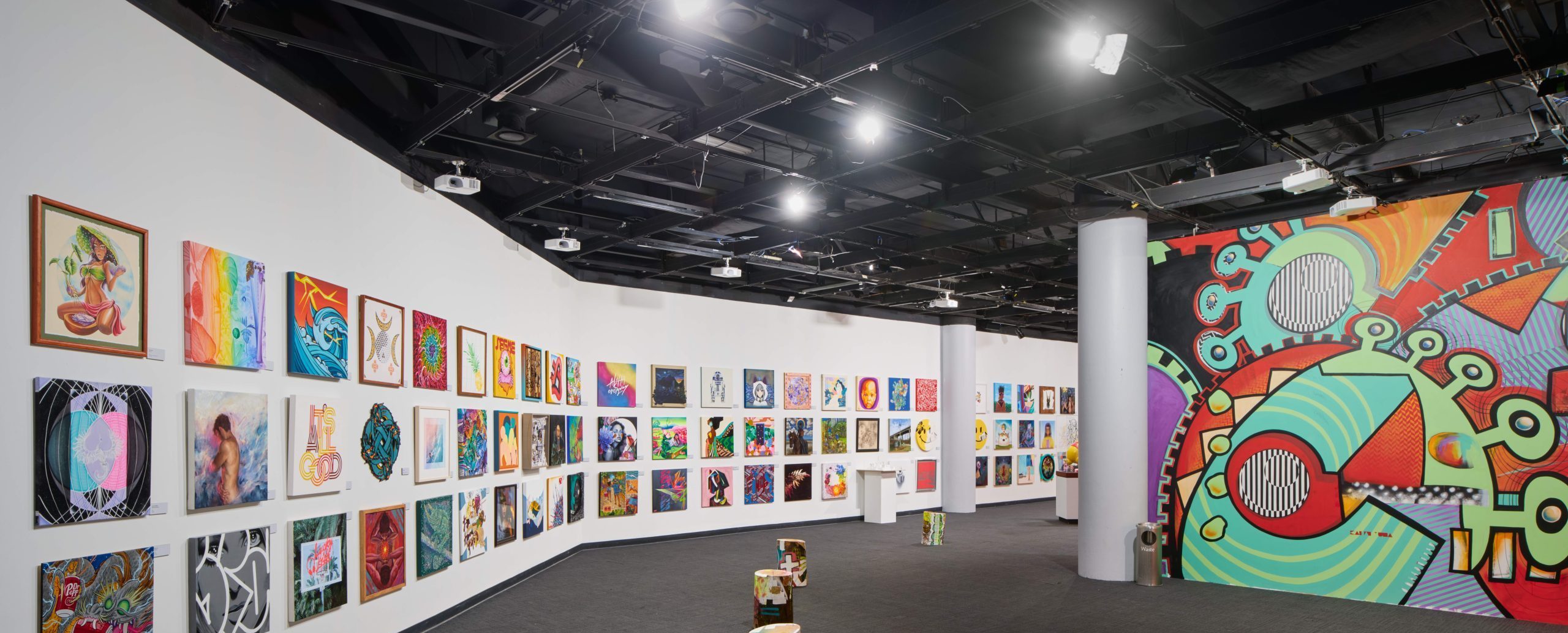 A long gallery wall hung with rows of colorful paintings beside a large abstract mural, under an exposed black ceiling with spotlights.