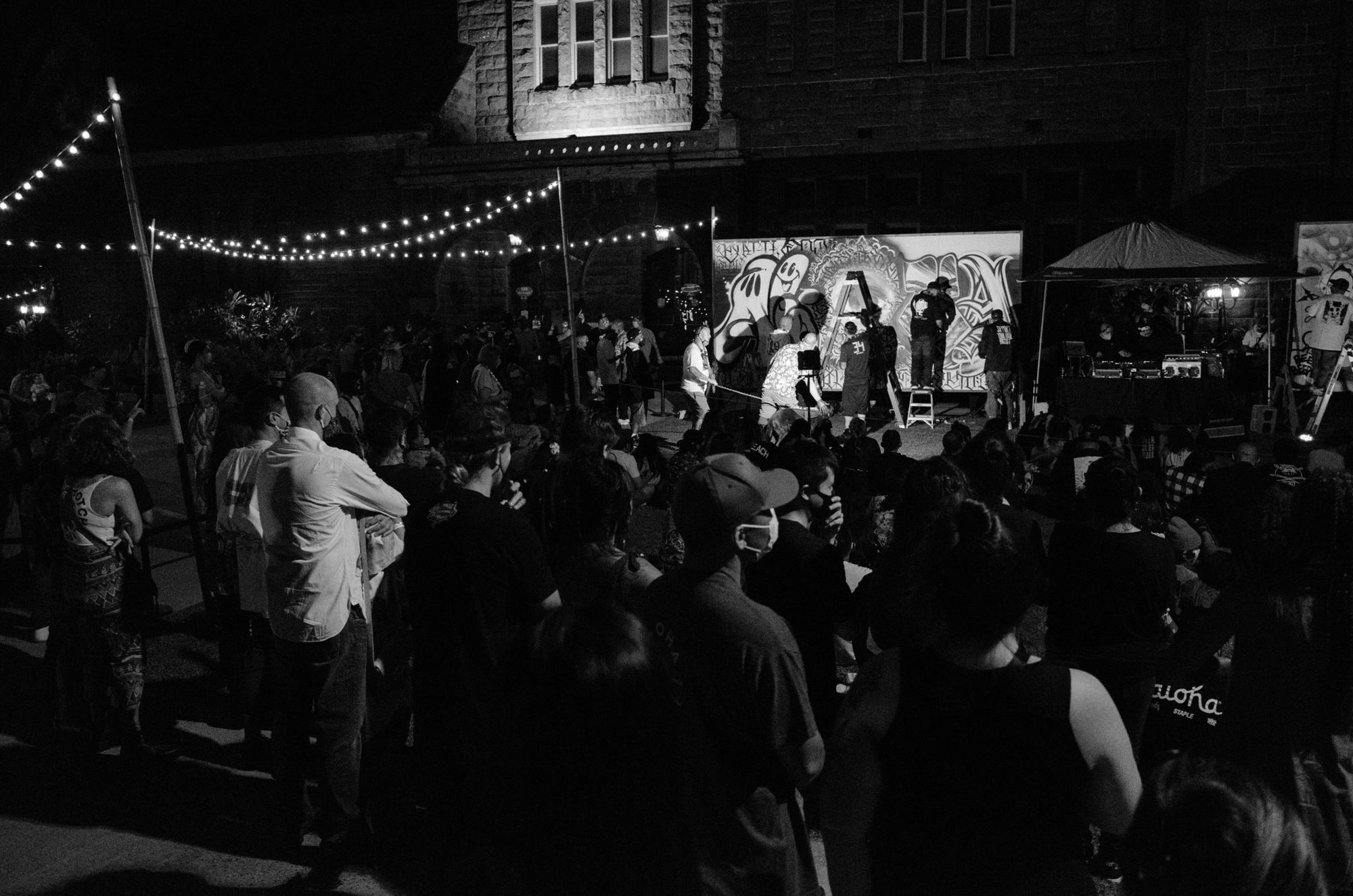At night under string lights, a crowd watches artists paint a large mural panel on the lawn in front of a lit stone building while a DJ plays.