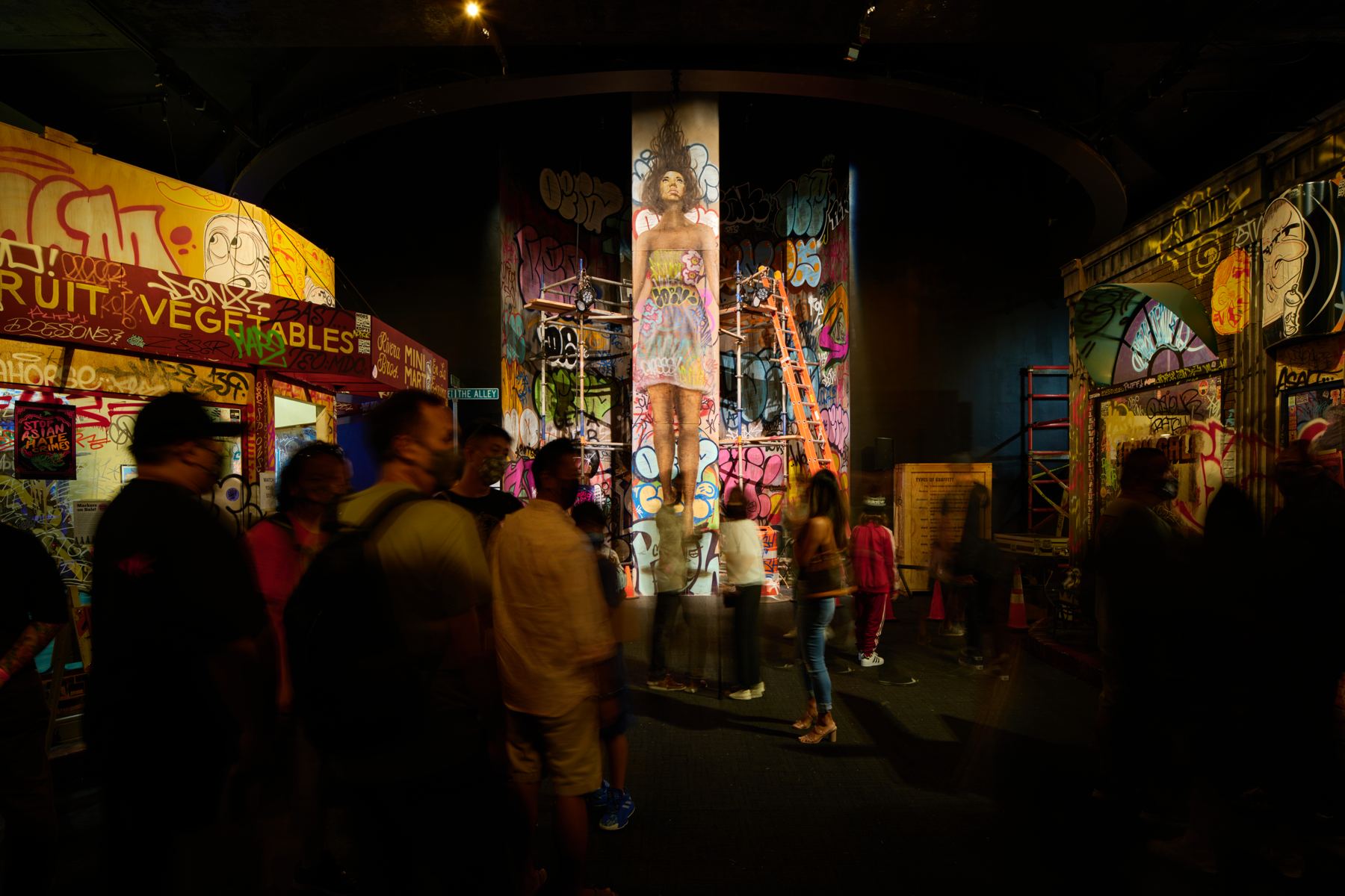 Visitors walk through a dim indoor streetscape lined with painted storefronts, murals and a mural-covered column.