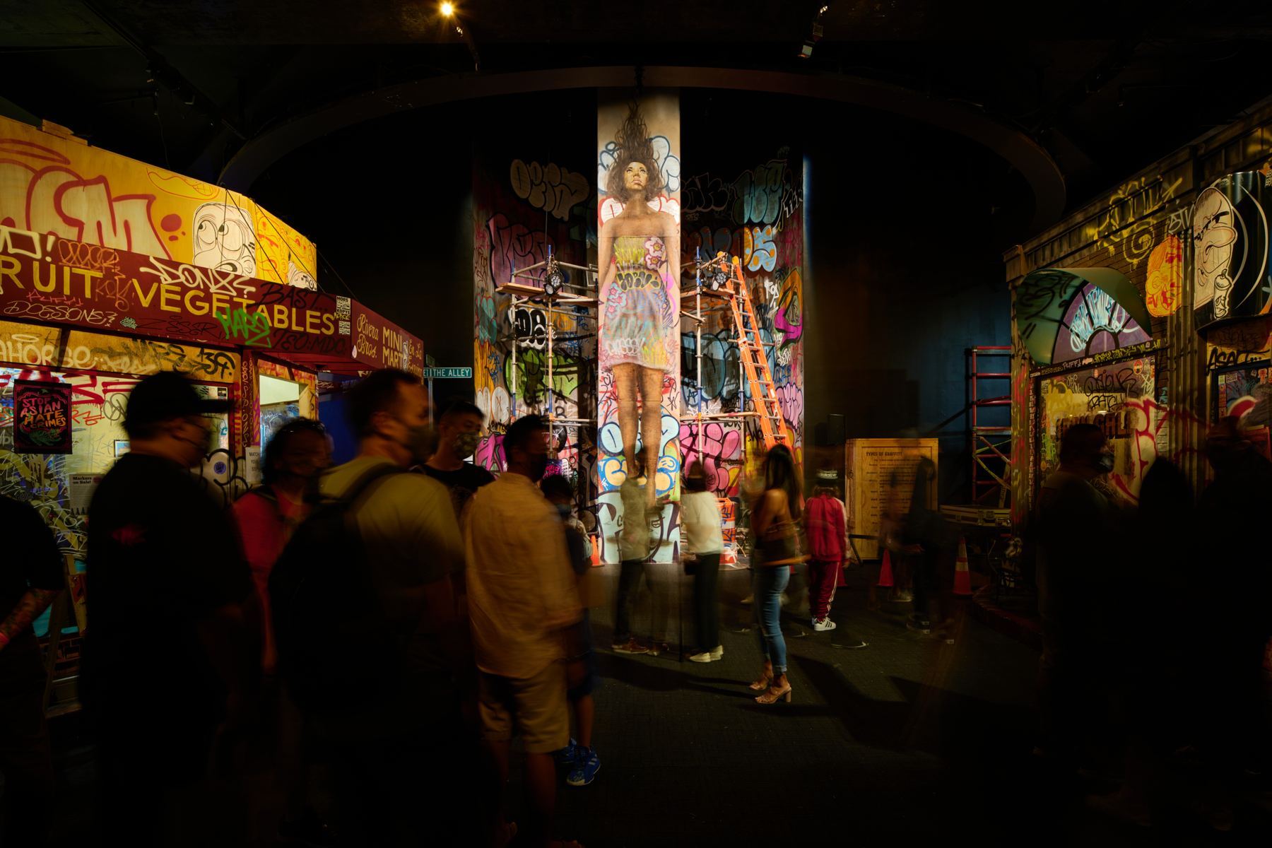 Visitors walk through a dim indoor streetscape of painted storefronts, murals and a mural-covered column.