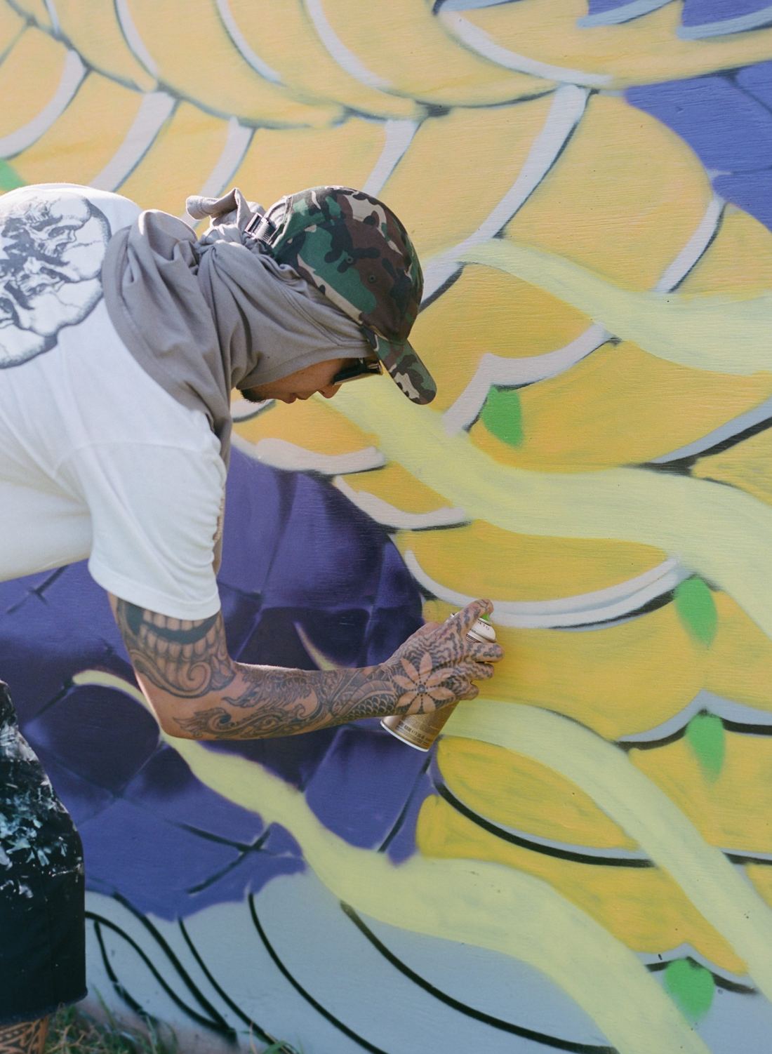 A tattooed artist in a camouflage cap and face covering sprays yellow paint onto a large mural.