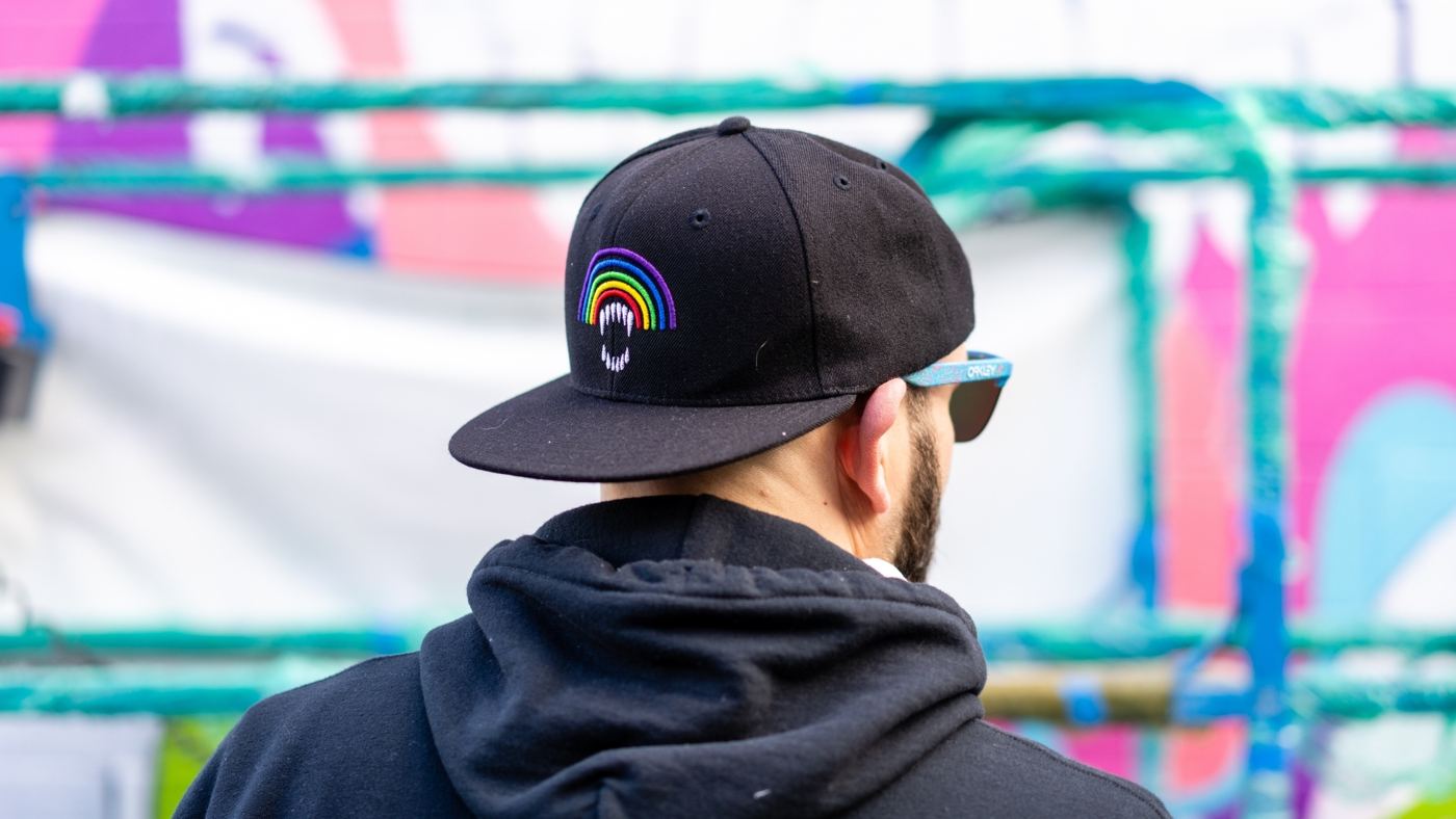 An artist in a black cap with a rainbow logo faces a brightly painted wall.