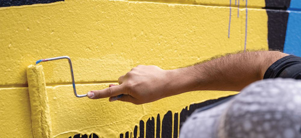 An arm rolling bright yellow paint over a mural wall.