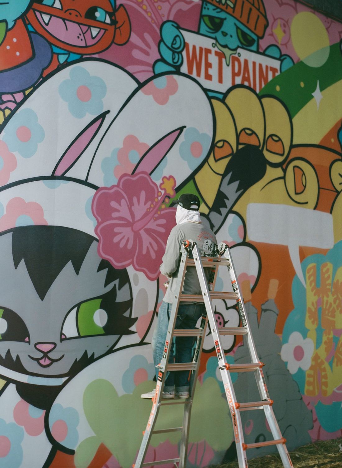 An artist on a ladder painting a large, colorful mural of cartoon characters.